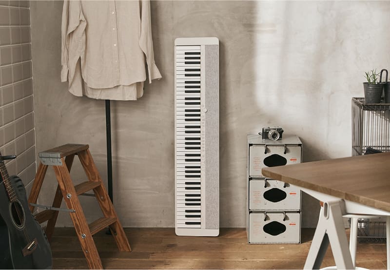 Un piano numérique blanc CT-S1-76WE est appuyé contre un mur gris, entre un portant à chemises et une petite commode