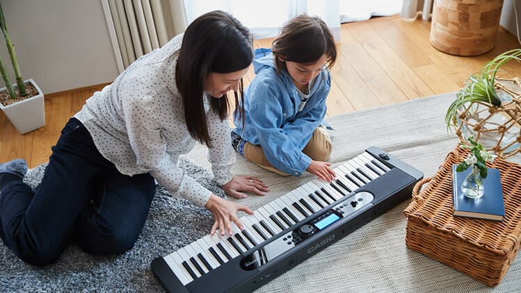 Une femme est accroupie sur le sol avec un enfant, ils jouent ensemble du piano numérique Casiotone CT-S400 posé devant eux sur le sol