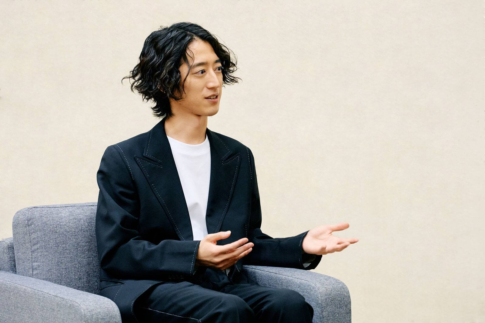 Hayato Sumino en costume noir, assis dans un fauteuil gris, en pleine conversation