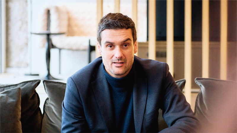 Vers la page Privia Magazine Voices d'Olivier Roset Olivier Roset est assis sur un canapé pendant l'interview.