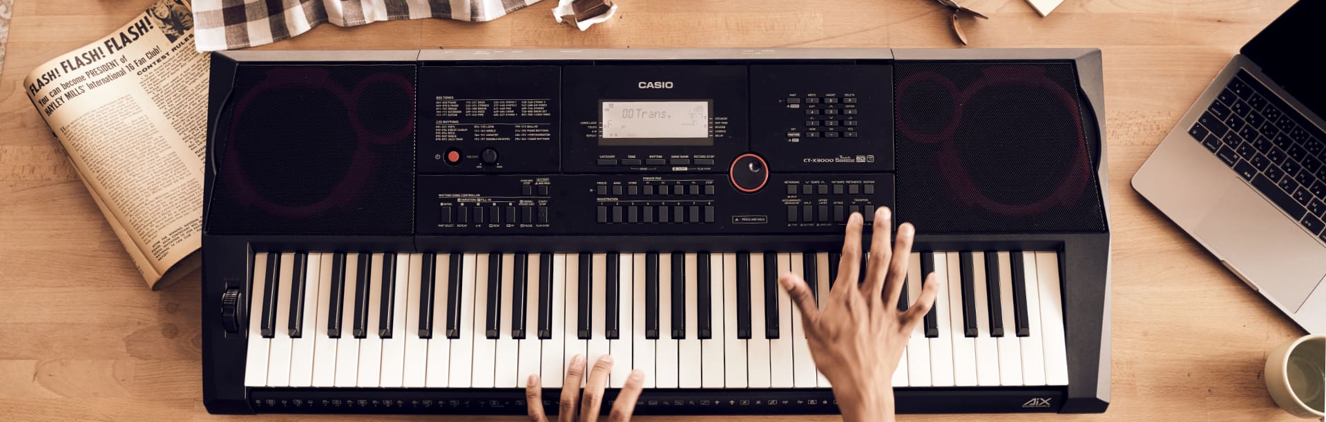 Le clavier noir CT-X avec de grands baffles et de nombreuses possibilités de réglage, vu d'en haut sur une table, à côté duquel se trouvent un journal et un ordinateur portable déplié