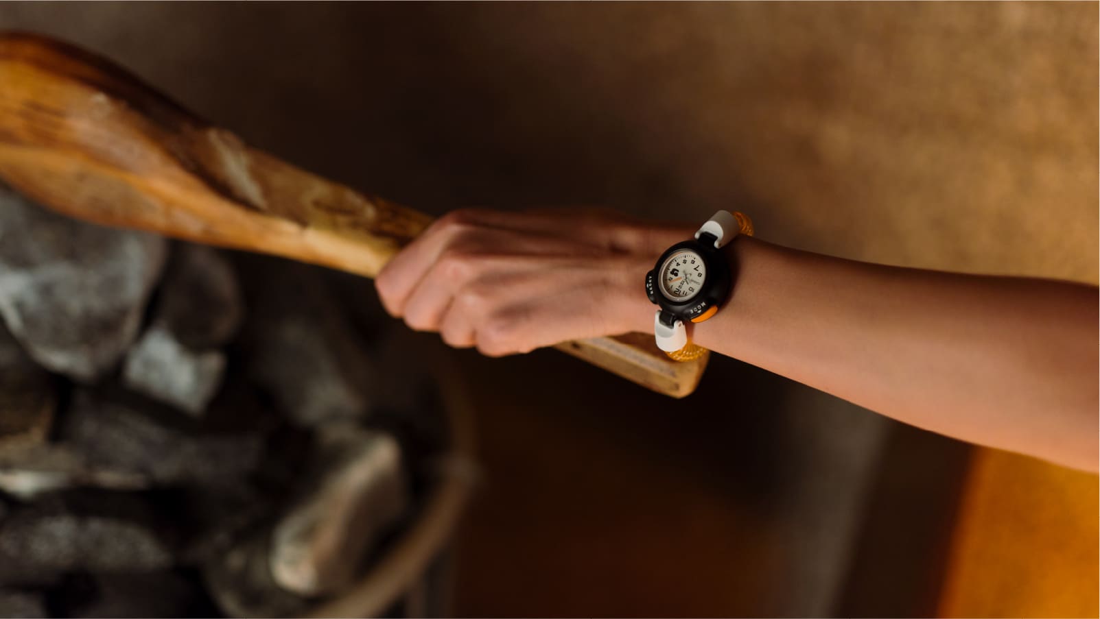 Gros plan sur le poignet d'une personne qui est dans un sauna. Elle verse de l’eau sur des pierres chaudes et porte la montre CASIO Sauna Watch noire et orange au poignet.