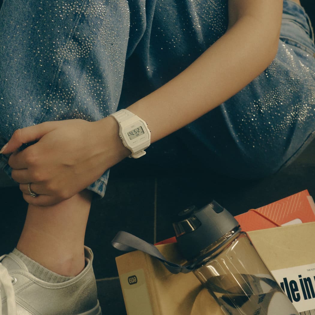 Un modèle féminin porte la F91WB-7AEF autour du poignet, à côté d'elle se trouve une poche ouverte avec une bouteille d'eau, un magazine et un classeur à l'intérieur. La montre CASIO TIMELESS COLLECTION est entièrement blanche et possède un affichage numérique de l'heure.