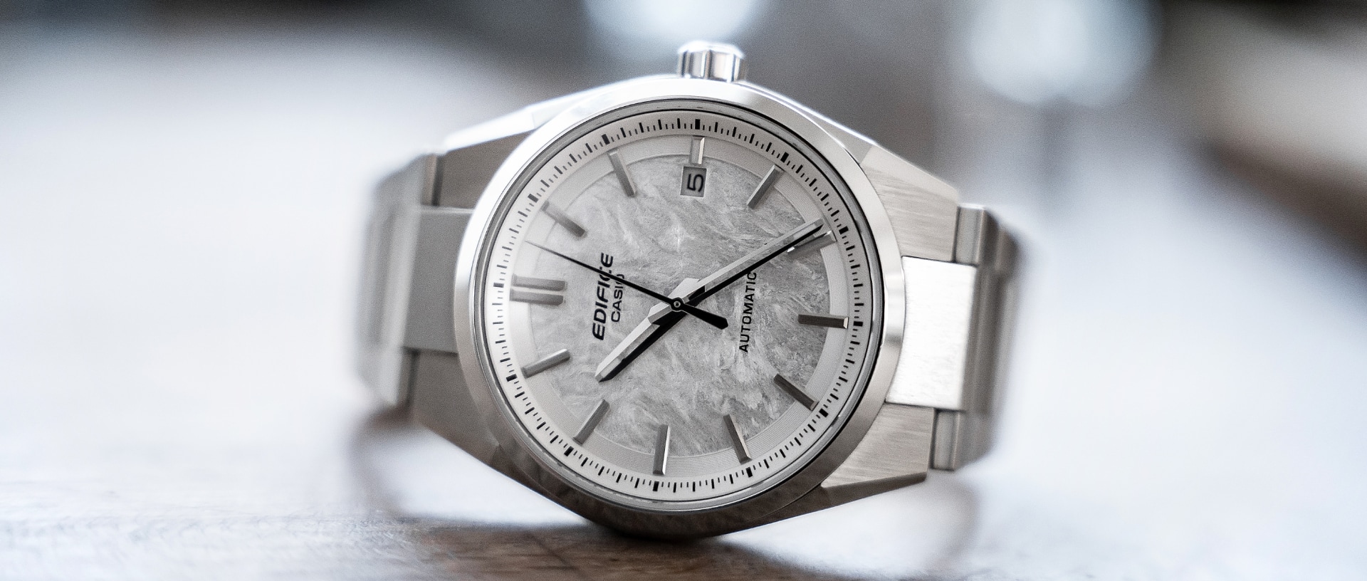 La montre automatique argentée EFK-110 d'EDIFICE avec un cadran texturé et des aiguilles argentées sur fond gris clair avec un motif représentant le mécanisme de l'horloge.