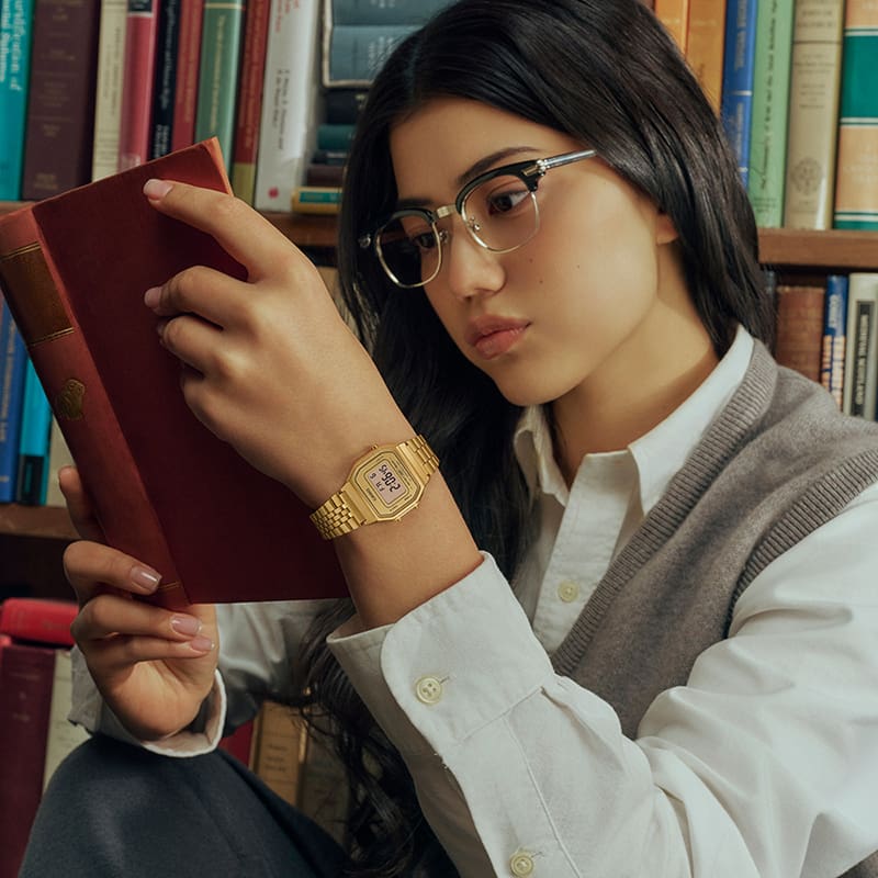 Une femme brune tient un livre dans ses mains. Elle porte des lunettes et la montre numérique CASIO Vintage Collection LA680WGA-9.