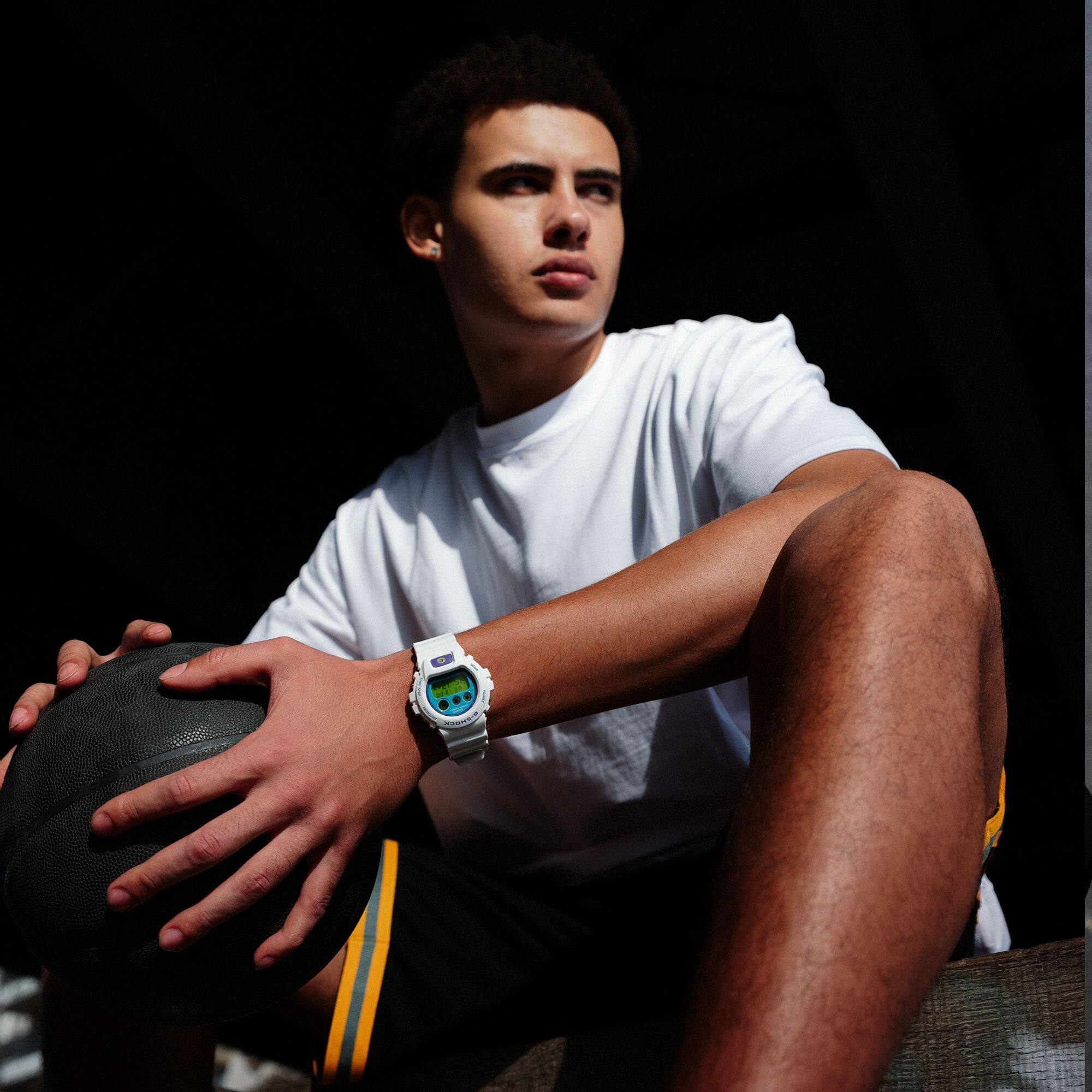 Un jeune homme est assis par terre avec un ballon de basket, il porte la DW-6900RCS-7 G-Shock blanche au poignet. L'image renvoie à la page du produit.