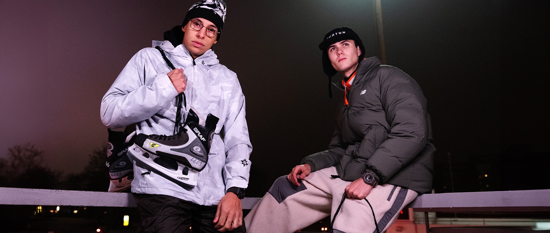 Deux hommes en vestes d'hiver, l'un portant des patins à glace sur l'épaule, l'autre assis sur une barrière. Tous deux portent au poignet des montres-bracelets G-Shock de la série GA-B2100CT.