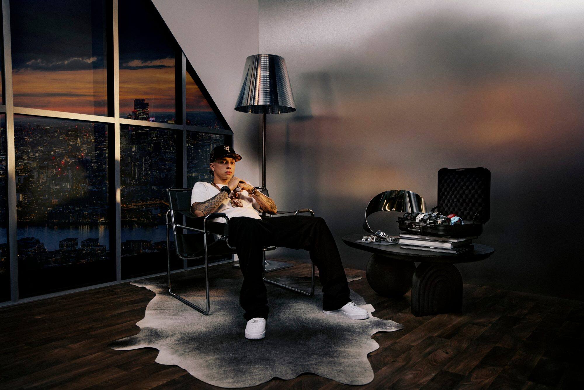 Central cee si rilassa su una sedia, indossa un G-Shock e ha una valigia piena di orologi sulla scrivania accanto a lui.