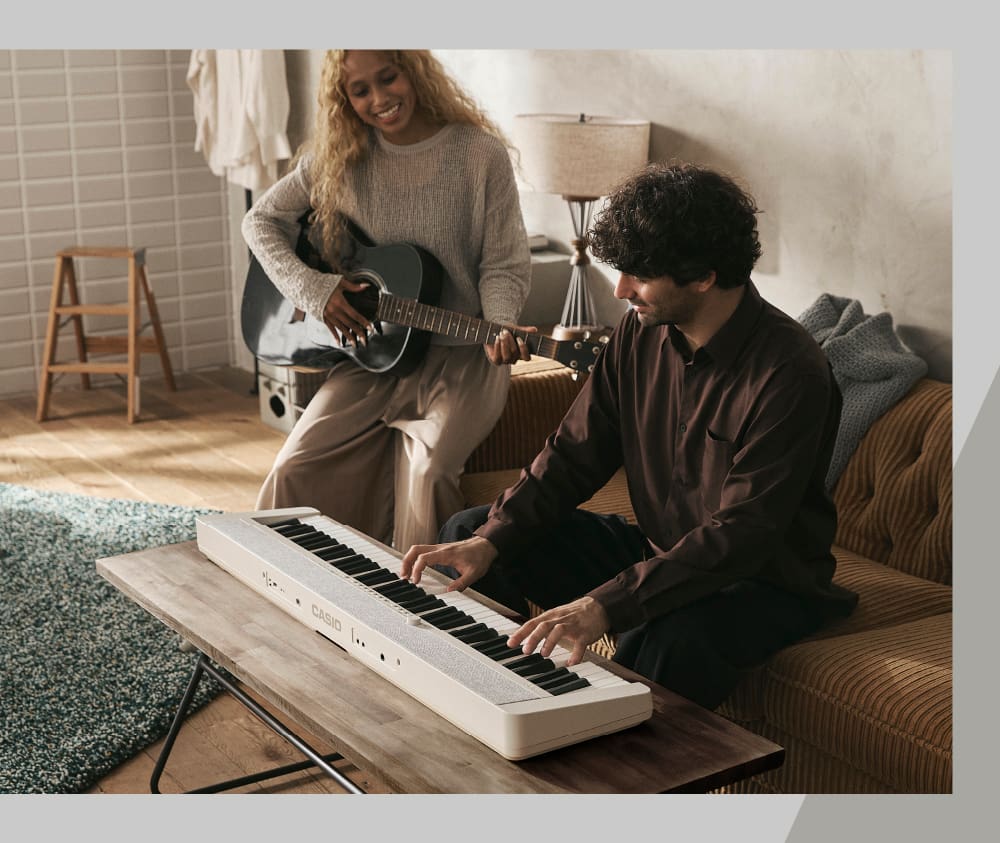 Un uomo è seduto su un divano e suona il pianoforte digitale Casiotone CT-S1-76 bianco che si trova sul tavolo del soggiorno di fronte a lui. Una donna lo accompagna alla chitarra