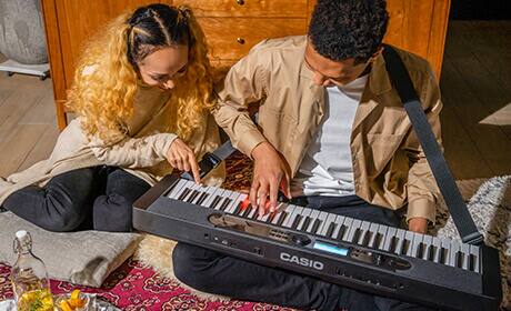 Una giovane donna e un giovane uomo sono accovacciati insieme sul pavimento e suonano insieme il pianoforte digitale Casiotone LK-S450, che è posizionato sulle ginocchia dell'uomo