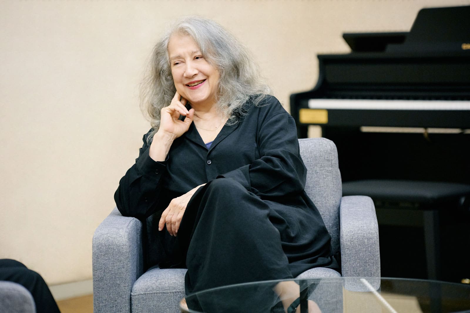 Martha Argerich seduta su un divano grigio, sullo sfondo si vede un pianoforte nero.