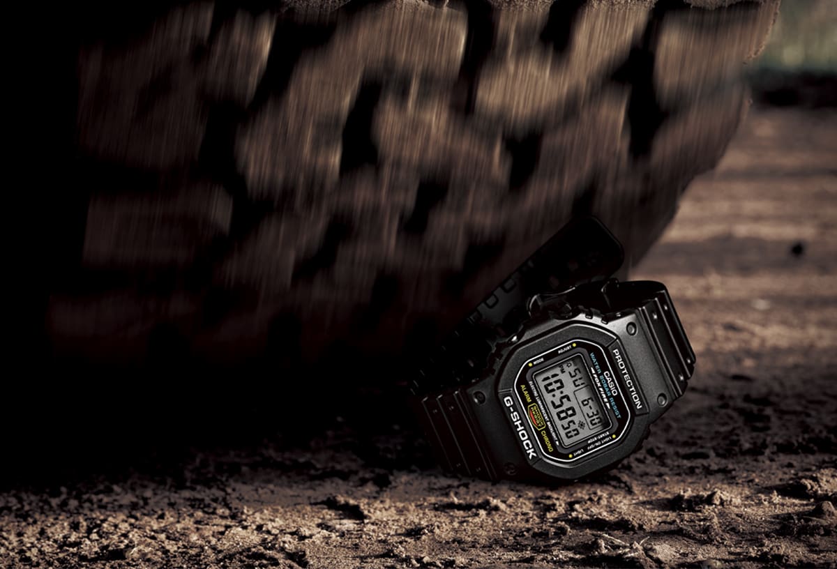Il G-SHOCK DW-5600 nero sul pavimento, dietro di esso un grande pneumatico che sta per passare sopra l'orologio.