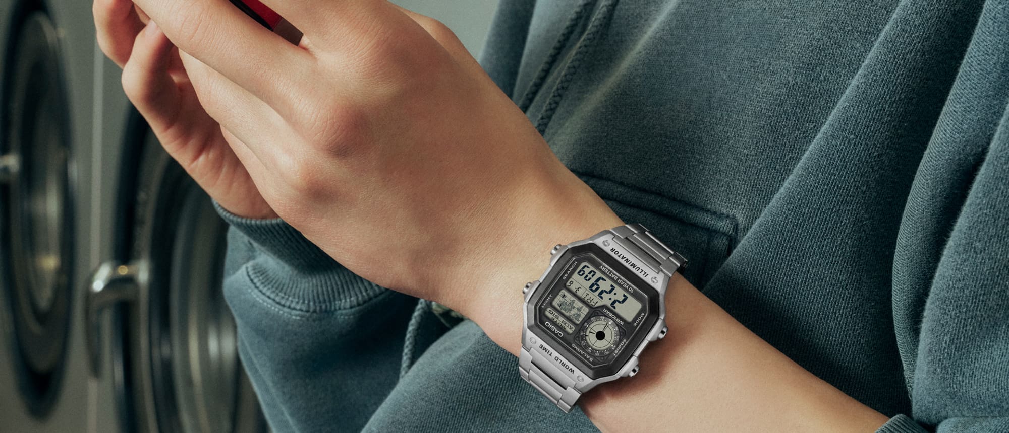 Un uomo tiene in mano il telefono indossando l’orologio digitale argento AE-1200WHD-1AV di Casio.