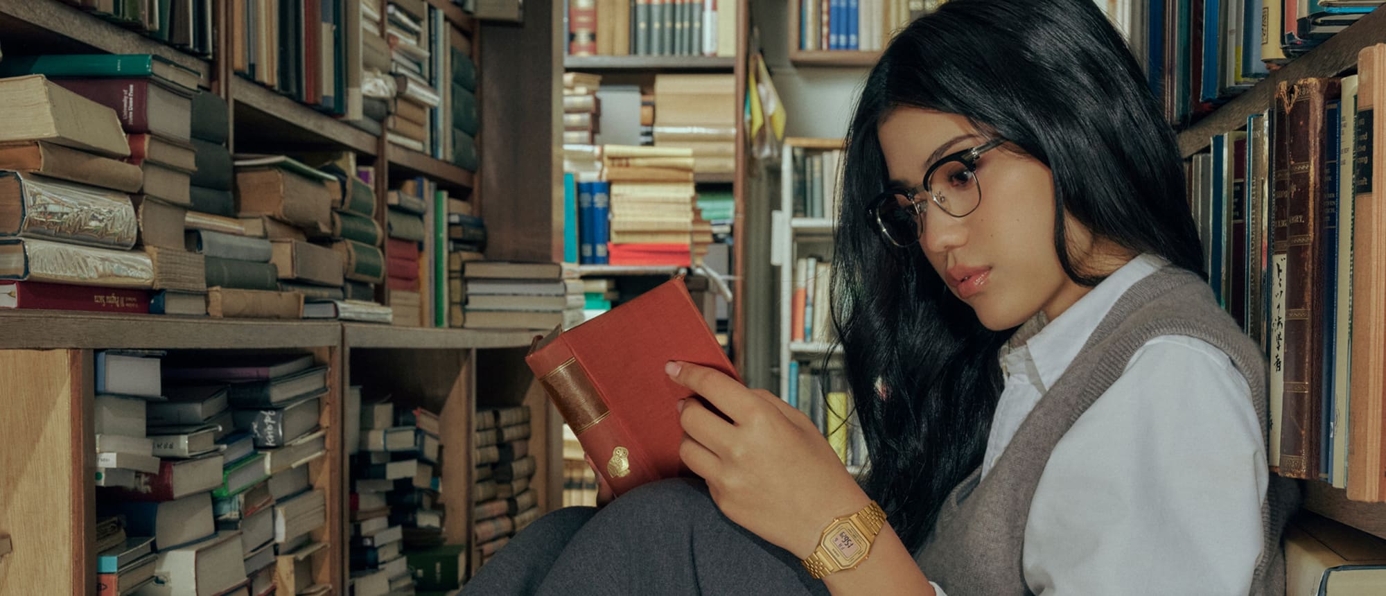Una donna dai capelli castani e gli occhiali legge un libro in biblioteca indossando l’orologio digitale dorato LA680WGA-9 della Collezione Casio Vintage.