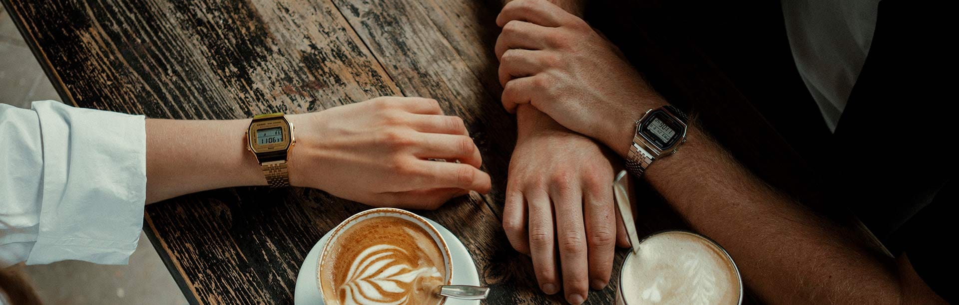 Due polsi che indossano gli ultimi orologi CASIO VINTAGE COLLECTION ABL-100 con cinturini color argento e oro giacciono su una scrivania di legno accanto a specialità di caffè.
