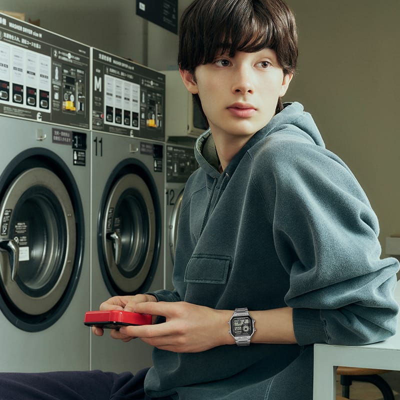  白いシャツを着た男性が、スクエア型のデジタルディスプレイとシルバーカラーのストラップが付いたCASIOタイムレスコレクションの腕時計を着け、一眼レフカメラのレンズを通して見ている。