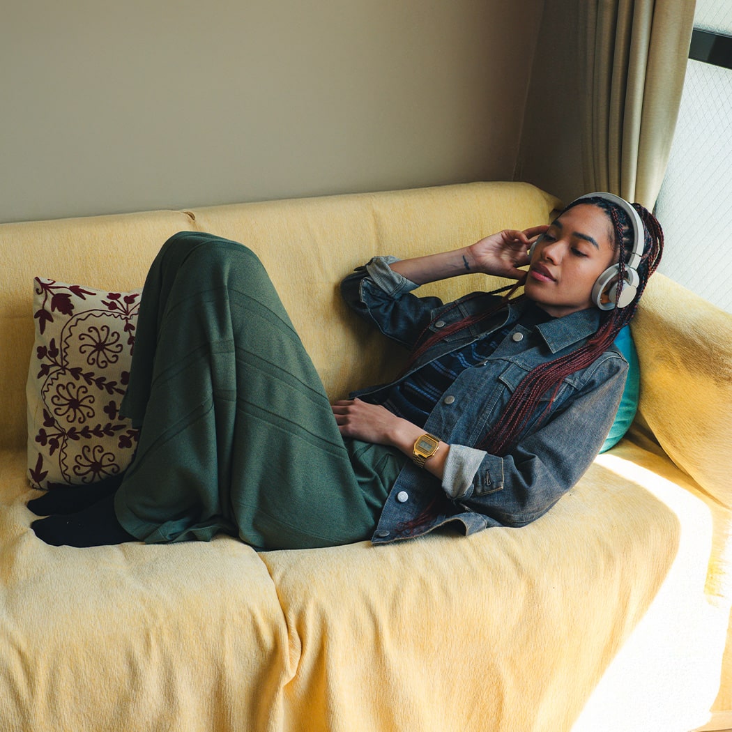 A woman sits on a yellow couch wearing denim clothes, grey headphones and the gold A168WG wristwatch from CASIO