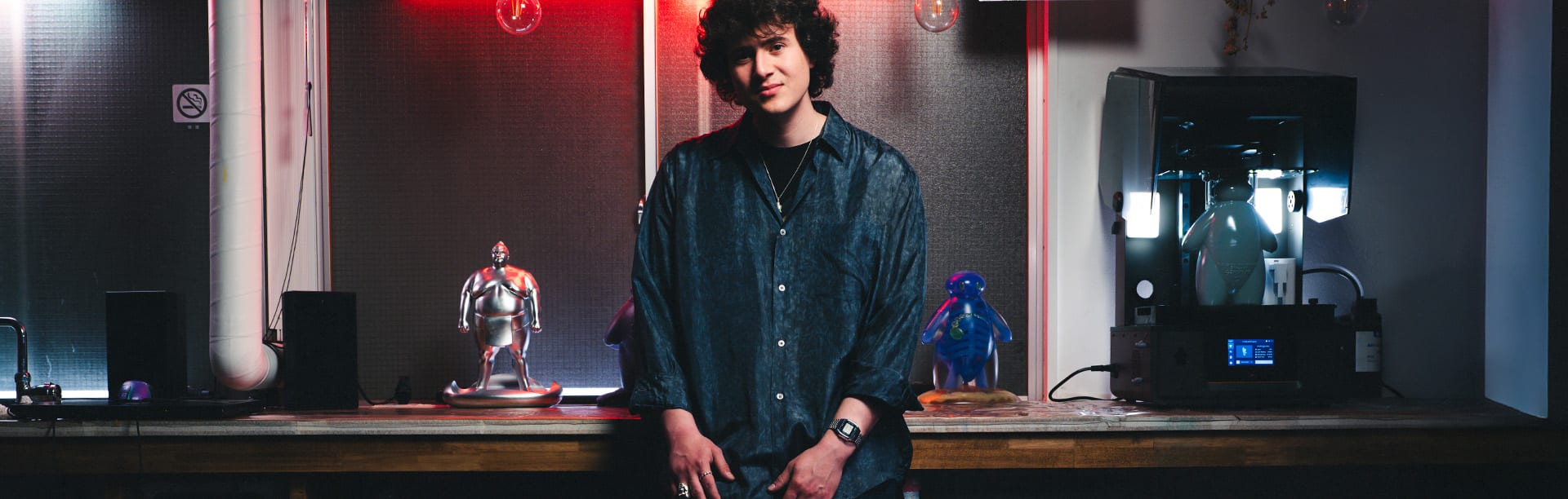 A man with curly hair and a dark shirt wears the silver-colored wristwatch ABL-100WE-1A on his wrist and stands in front of a workbench with a 3D printer