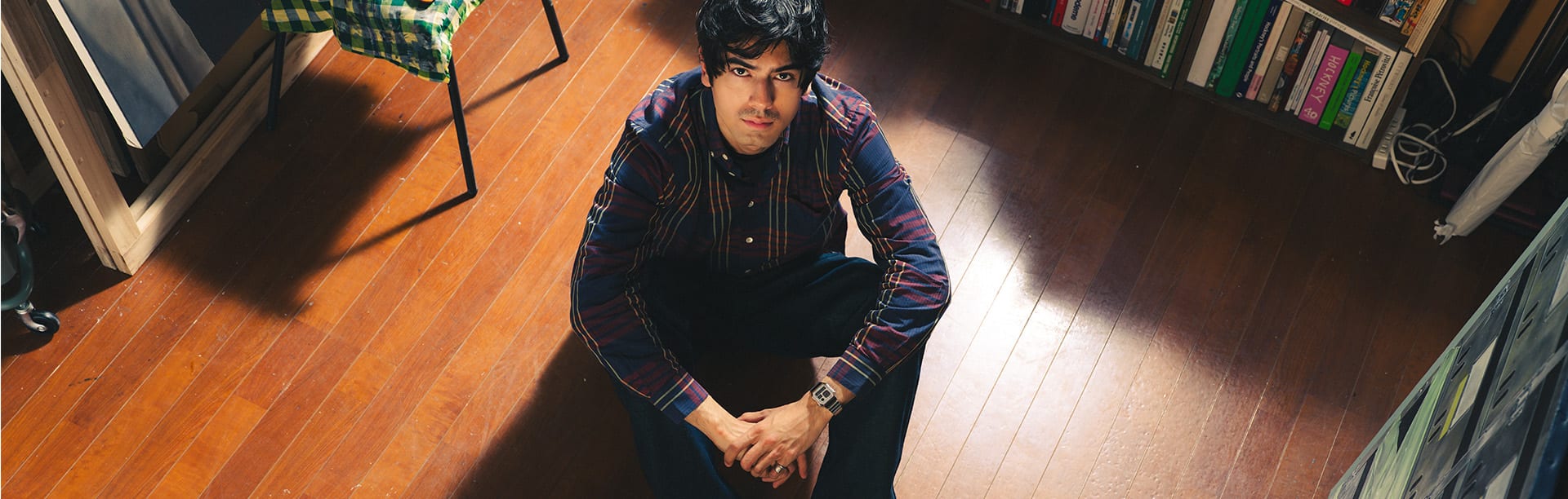 A man in a checkered shirt sits on a wooden floor in an apartment and looks up at the camera, wearing the silver-colored digital watch A130WE-7A from CASIO on his wrist
