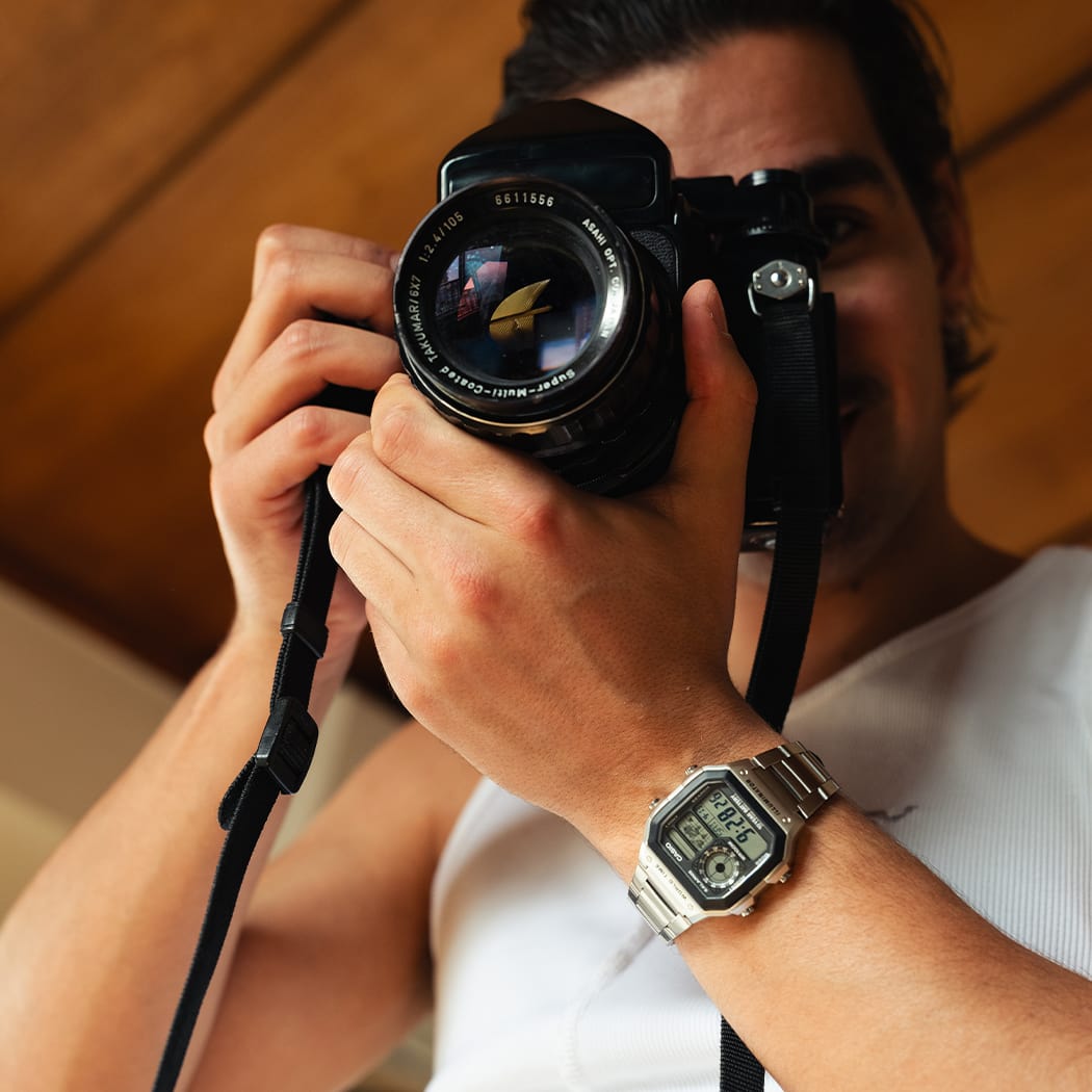 Un hombre con una camisa blanca que usa el reloj CASIO Timeless Collection con pantalla digital cuadrada y correa de color plata y mira a través del lente de una cámara réflex