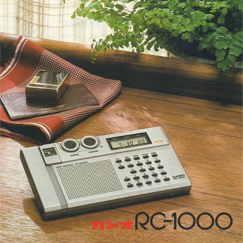 Een rechthoekige radio in zilvergrijs met een ingebouwde rekenmachine en een kleine klok op een houten bureau.