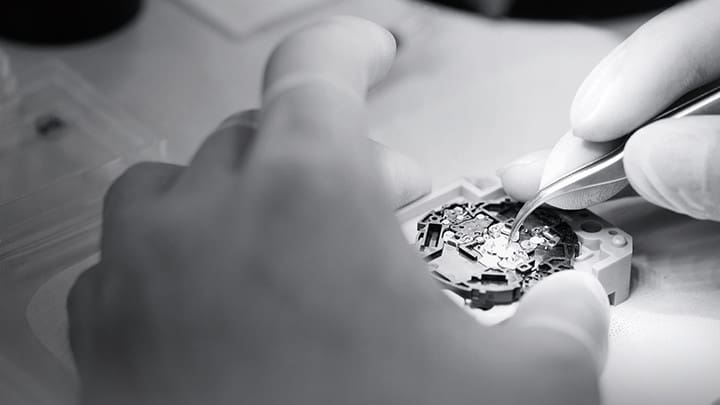 Close-up van de handen van een persoon die zorgvuldig een uurwerk in elkaar zet.