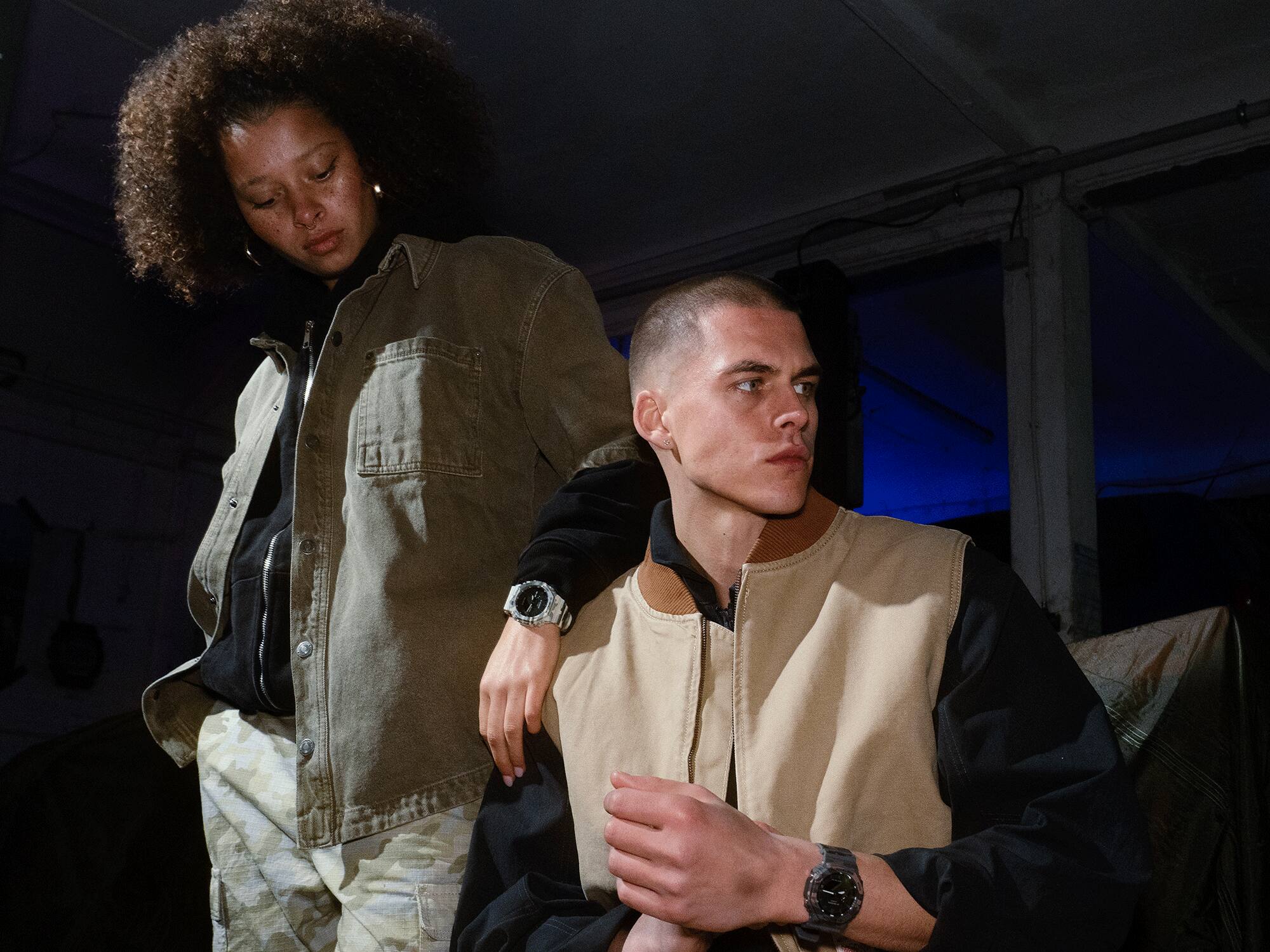 Een jonge vrouw leunt nonchalant met gebogen arm tegen een zittende jongeman. Beiden dragen vrijetijdskleding en een G-Shock GA-2100CM model om hun pols.