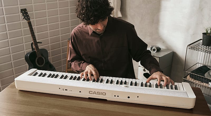 Een man zit aan een houten tafel en bespeelt de witte Casiotone CT-S1-76 digitale piano