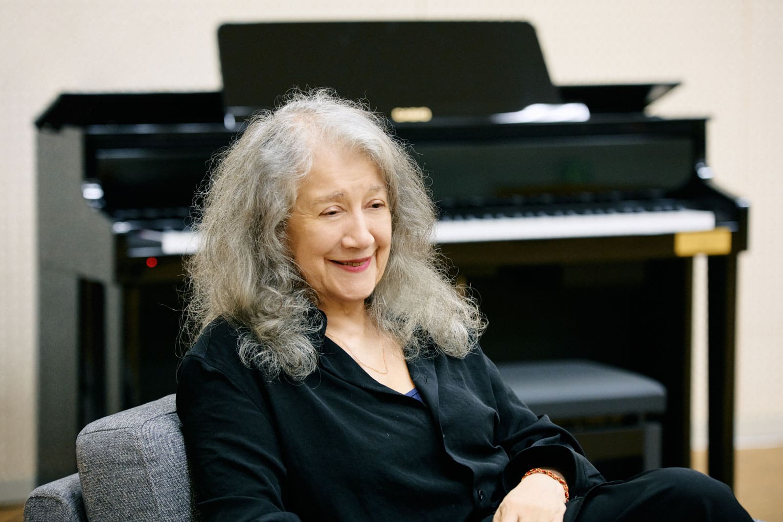 Martha Argerich zit op een grijze bank, op de achtergrond is een zwarte piano te zien.
