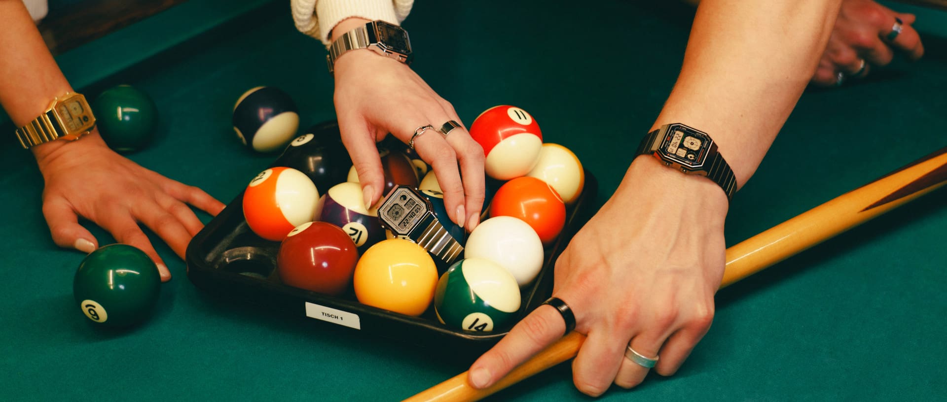 Drie handen reiken naar biljartballen en het digitale horloge A130 op een pooltafel, met de CASIO Vintage modellen A130 in verschillende kleuren.