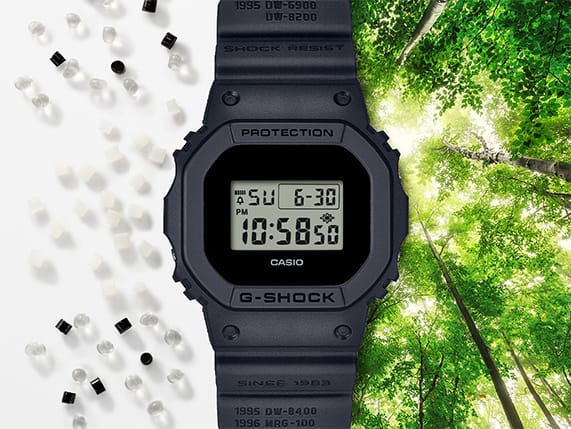 Een digitaal horloge uit de jubileumeditie tussen kunststofgranulaat op een witte ondergrond en het uitzicht op een licht bladerdak