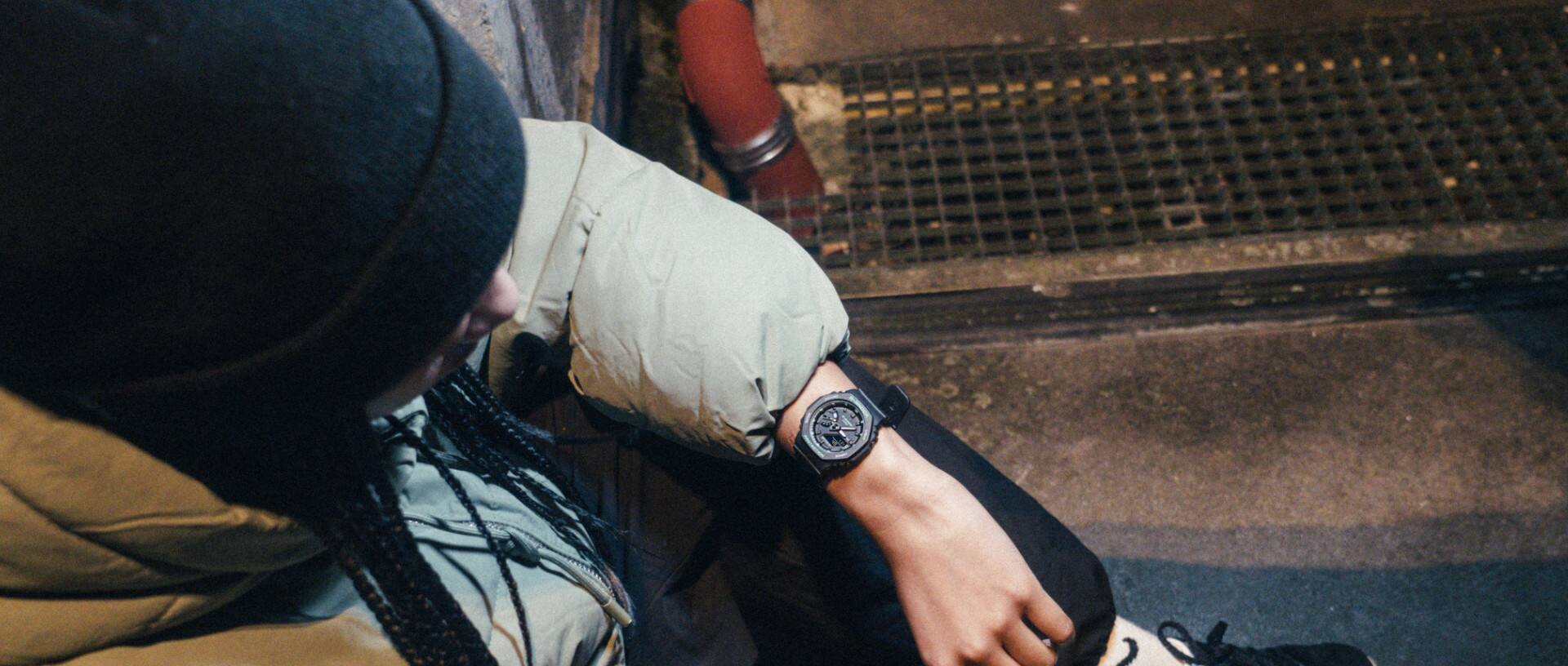Bovenaanzicht van iemand die in winterkleding op een stoeprand zit en naar zijn zwarte G-Shock GMA-S21000 horloge kijkt.