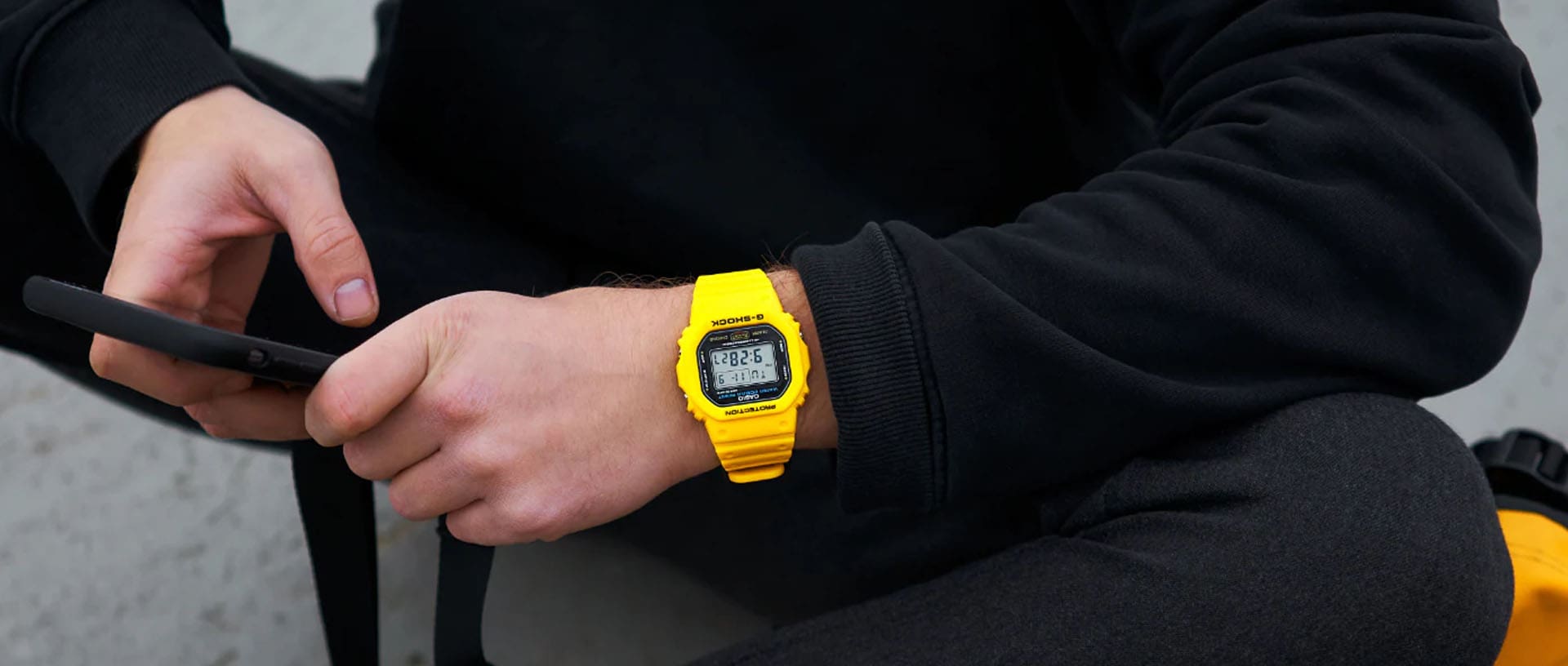 Een persoon in zwarte kleding gebruikt zijn smartphone en draagt een geel G-Shock digitaal horloge om zijn pols