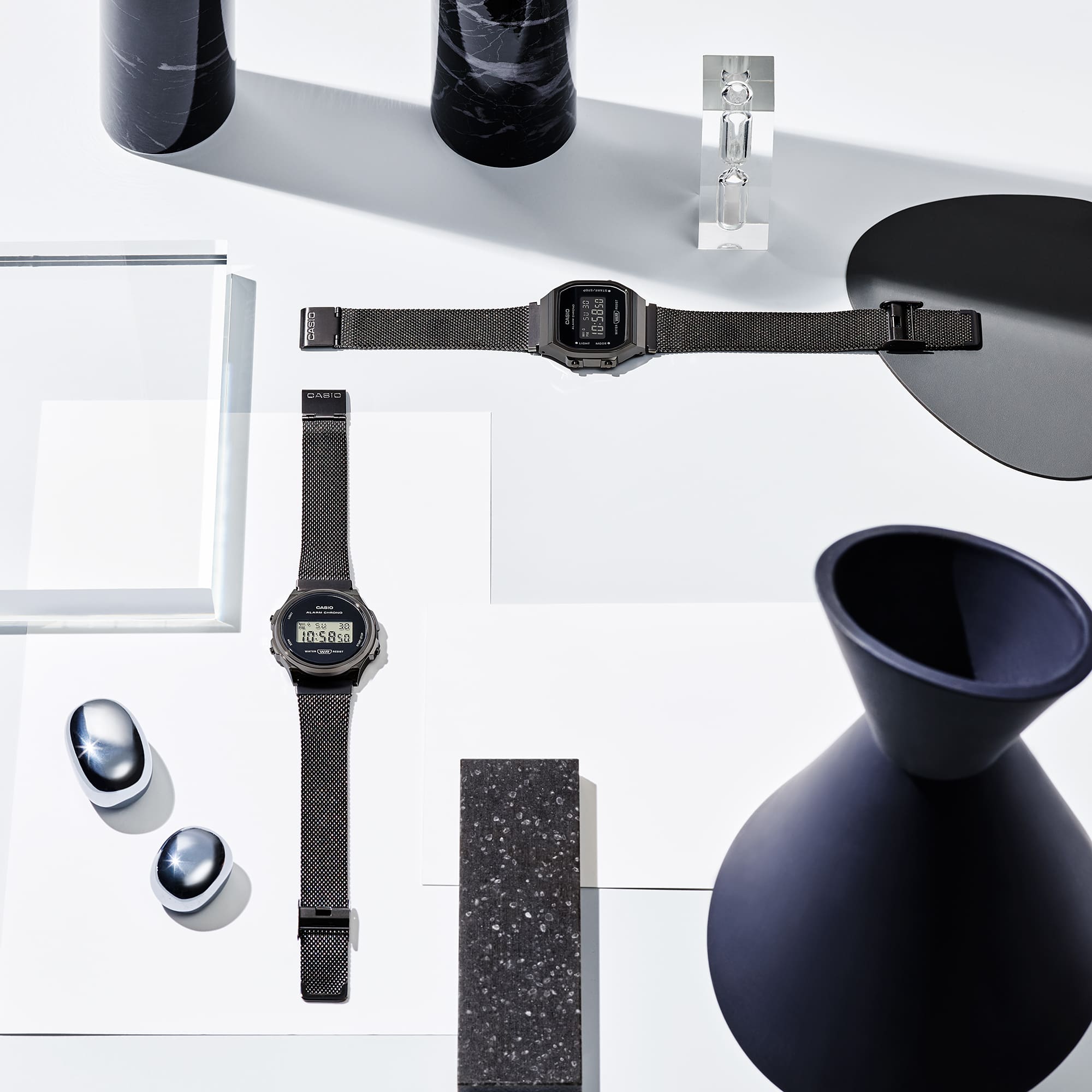 De zwarte digitale horloges A168WEMB-1B en A171WEMB-1A zijn tussen verschillende accessoires op een witte tafel gerangschikt. De afbeelding linkt naar A168WEMB-1B.