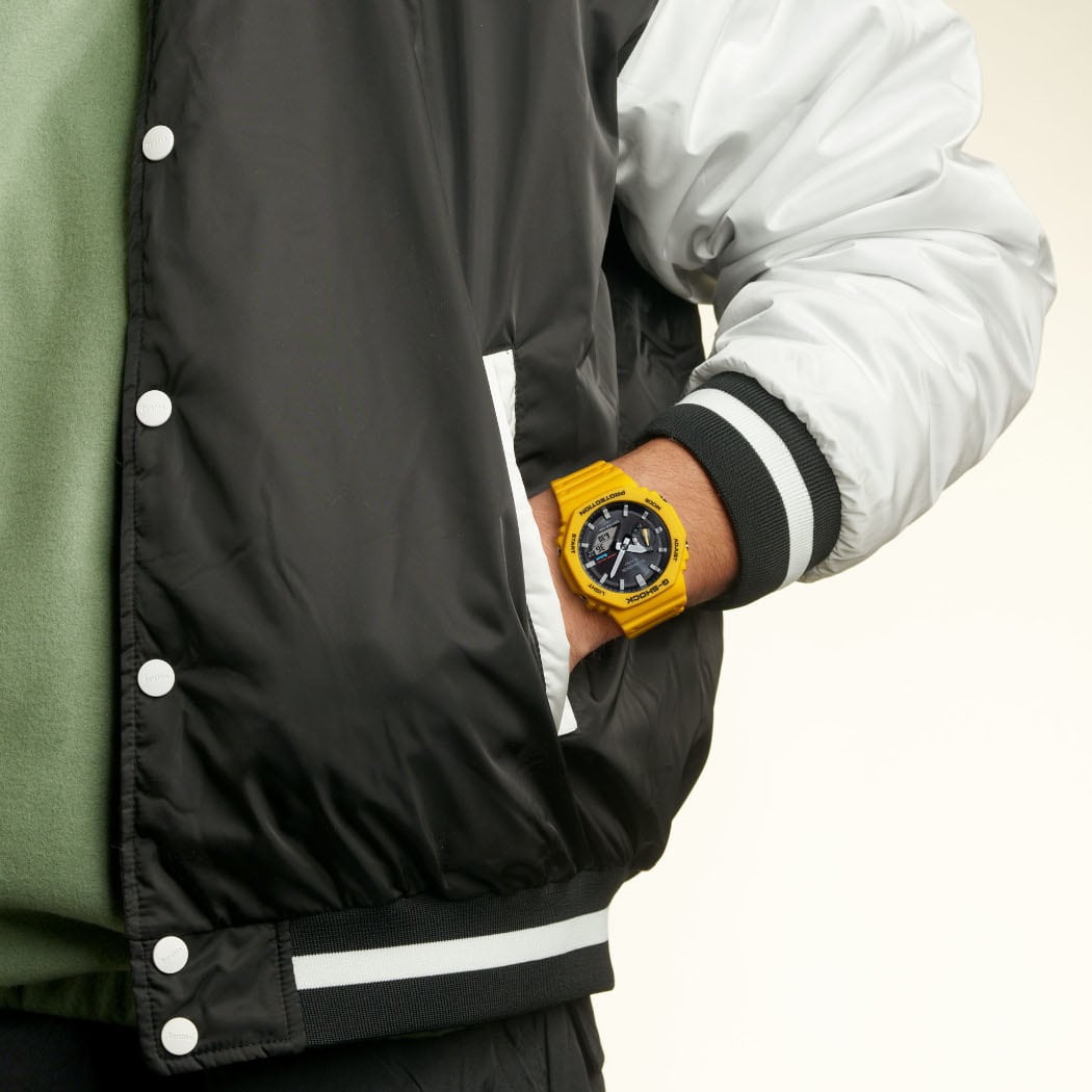 Iemand draagt een baseballjack met zijn hand in de jaszak, om de pols zit het Casio solar horloge GA-B2100C-9A met gele band en zwarte wijzerplaat