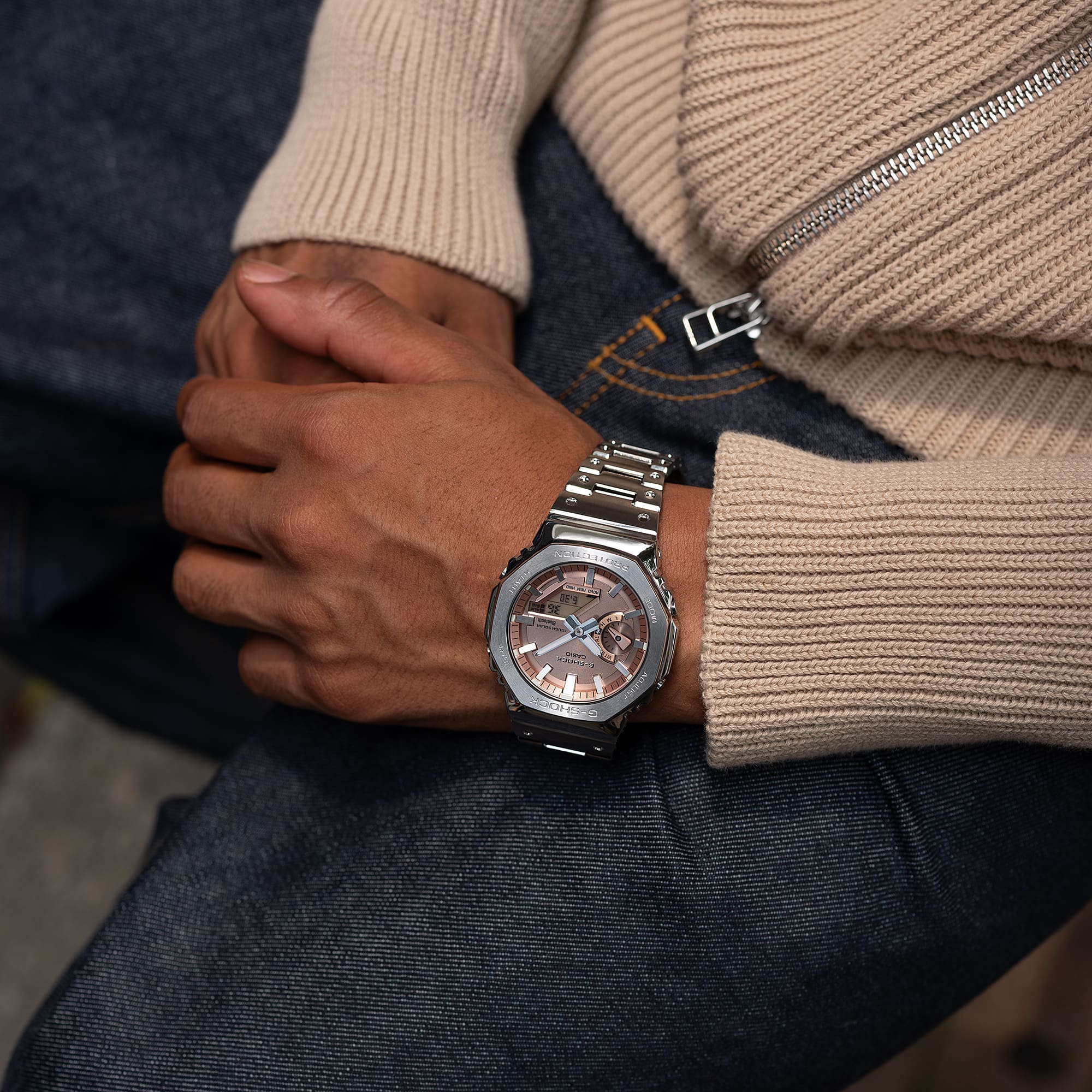 Het hybride polshorloge GM-B2100AD-5A van Casio G-Shock met zilverkleurige roestvrijstalen horlogeband en koperkleurige wijzerplaat om de pols van een man die een beige vest draagt