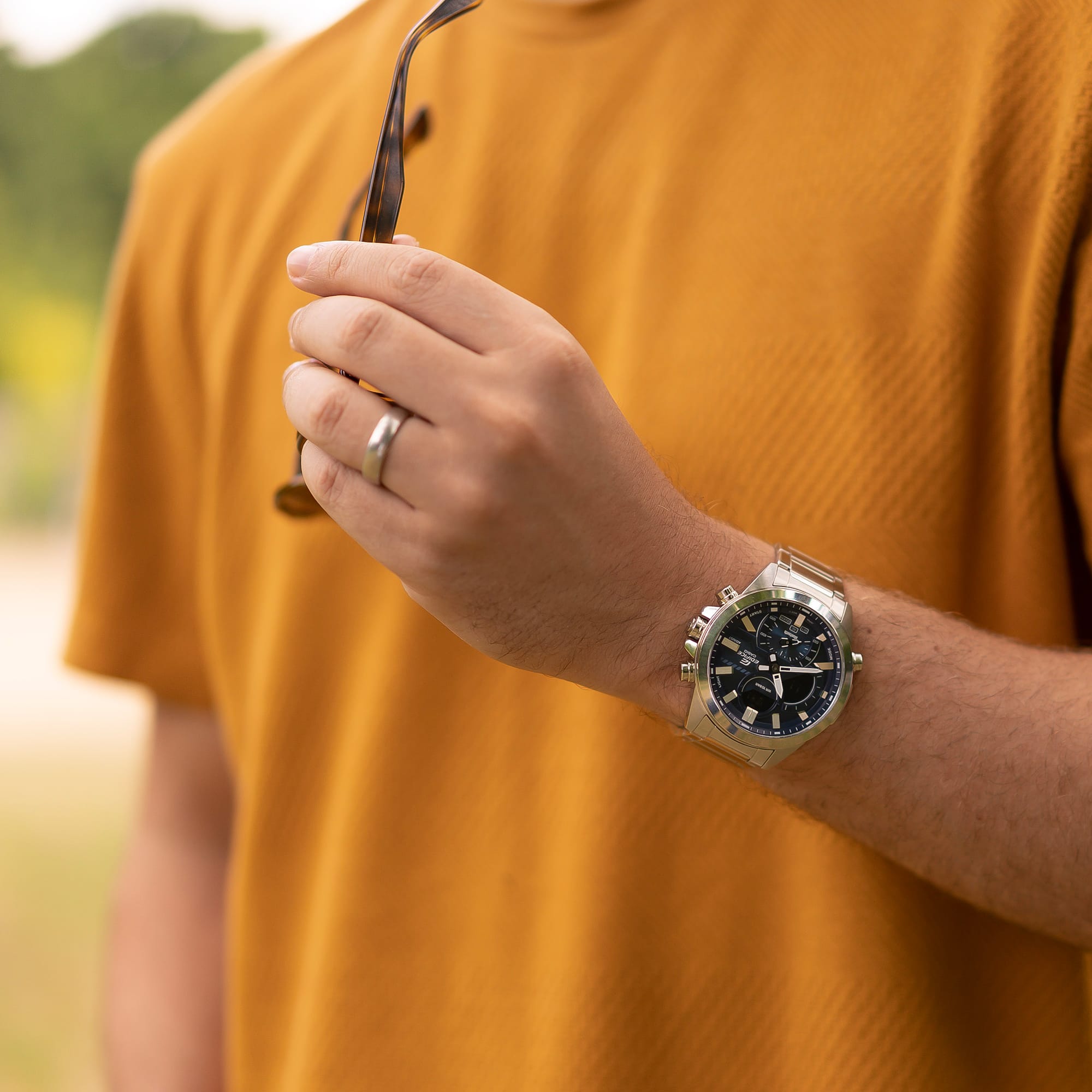De zilveren Casio Edifice ECB-30D-2A roestvrijstalen chronograaf met een blauwe, digitaal-analoge wijzerplaat om de pols van een man in een geel T-shirt