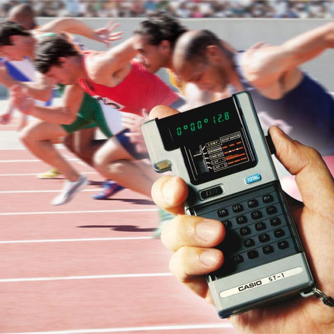 Uma mão cronometra o tempo com a calculadora-cronómetro ST-1; ao fundo, os corredores dão a partida. A imagem leva ao artigo sobre a ST-1.