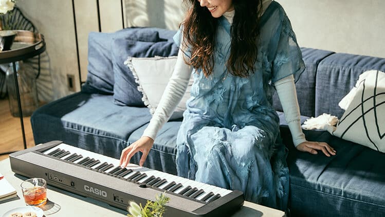 Uma jovem mulher com um vestido azul claro senta-se num sofá e toca o Casiotone CT-S1BK preto à sua frente na mesa da sala de estar