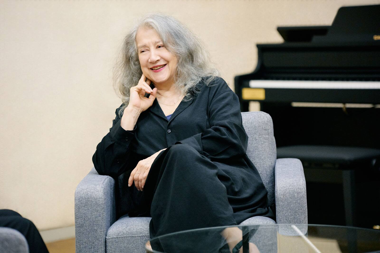 Martha Argerich está sentada num sofá cinzento, com um piano preto ao fundo.