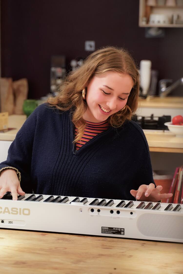 A estudante Antonie Weber está sentada à mesa da cozinha e toca o seu piano Privia branco. A imagem tem um link para a sua página Viver com Privia.