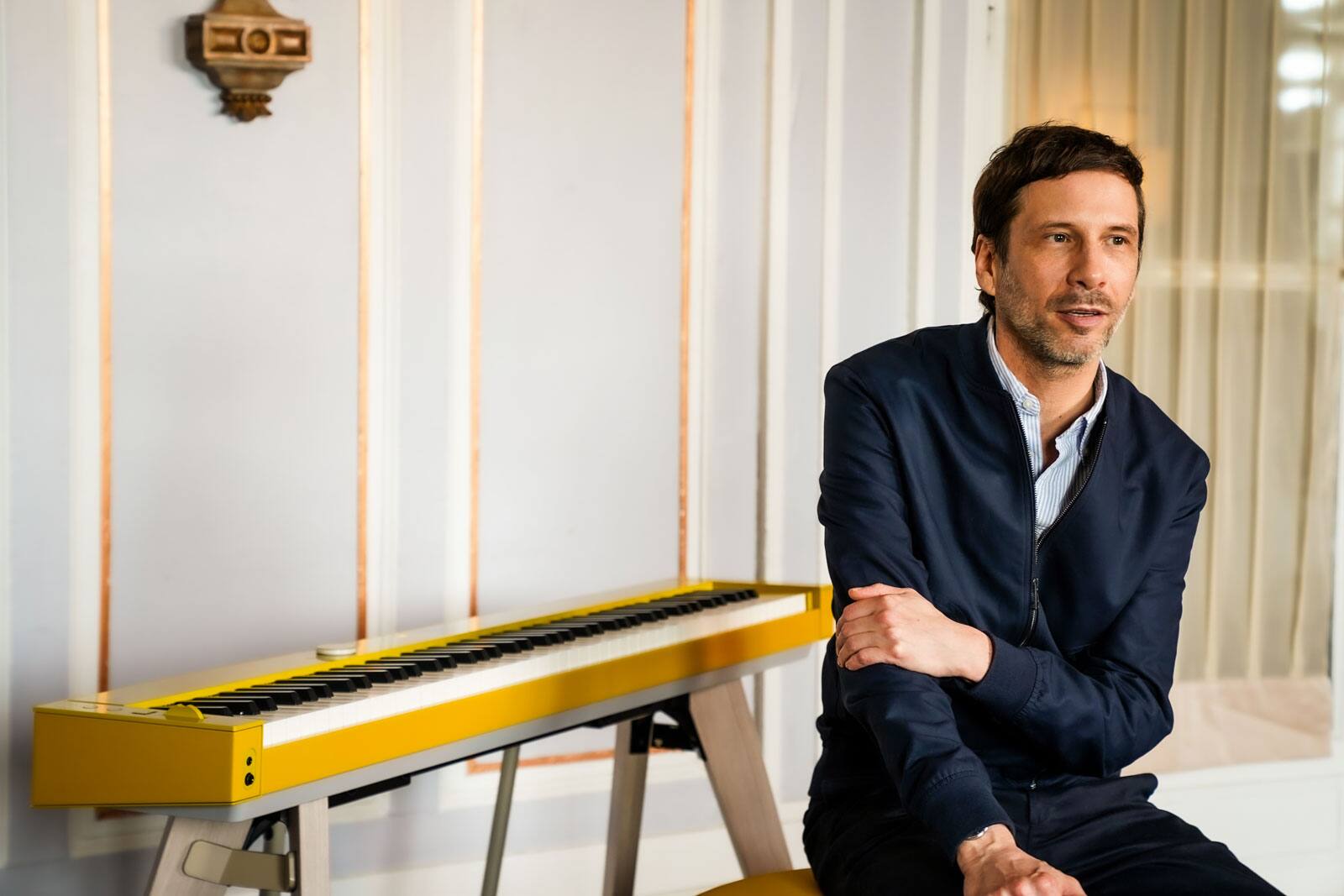 O cantor e compositor francês Alex Beaupain sentado num banco de piano ao lado do seu piano digital Privia PX-S7000HM amarelo