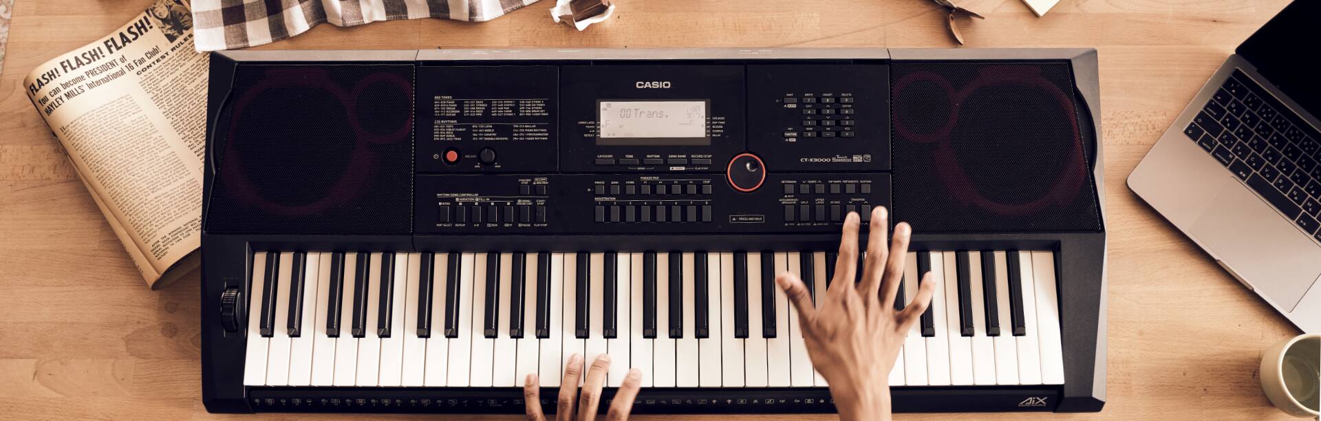 O teclado CT-X preto com altifalantes de grandes dimensões e muitas opções de configuração em cima de uma mesa, ao lado de um jornal e de um computador portátil aberto