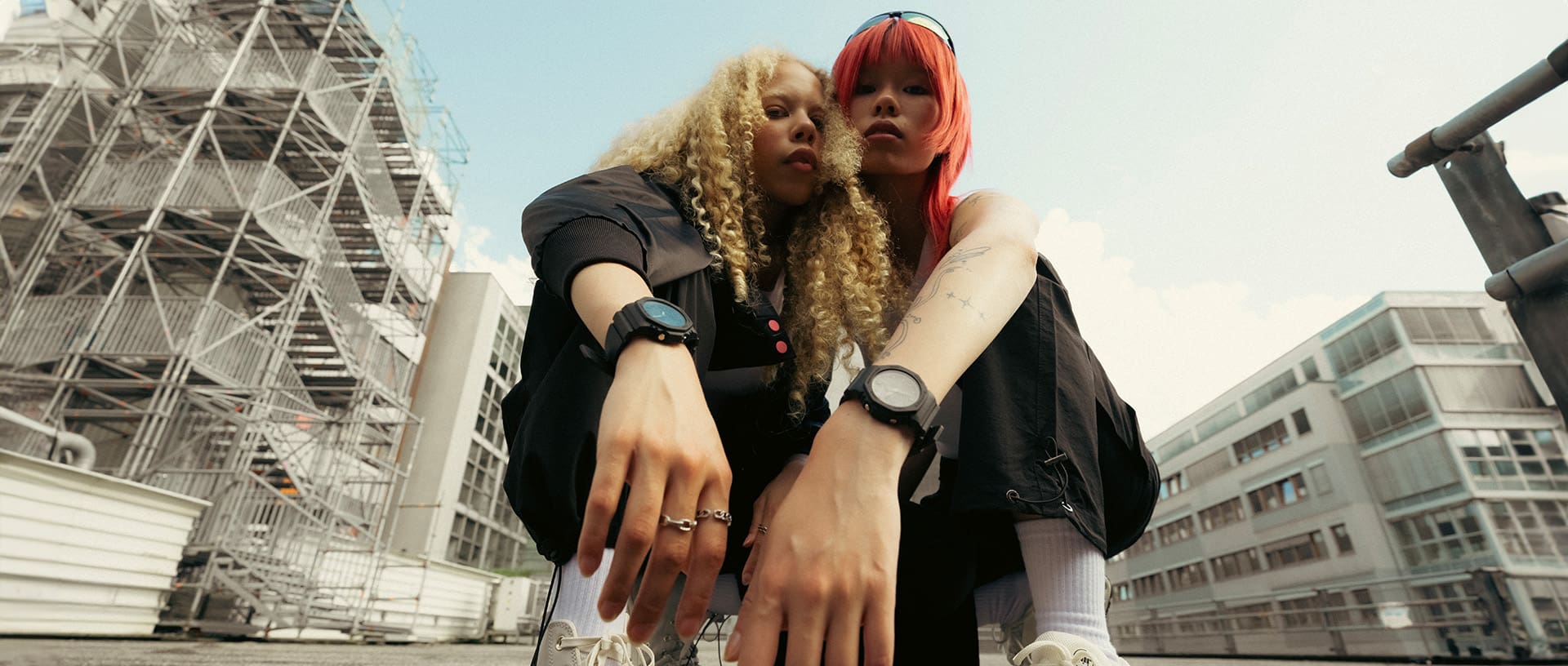 Duas mulheres agacham-se em frente a fachadas cinzentas, olham para a câmara e usam versões pretas do relógio digital analógico G-SHOCK “2100” nos pulsos