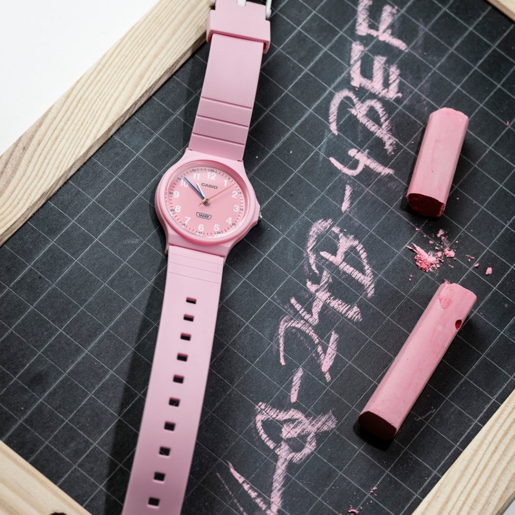 O relógio de pulso analógico LQ-24B com bracelete e mostrador cor-de-rosa está sobre uma pequena tábua. A designação completa do produto está escrita a giz.