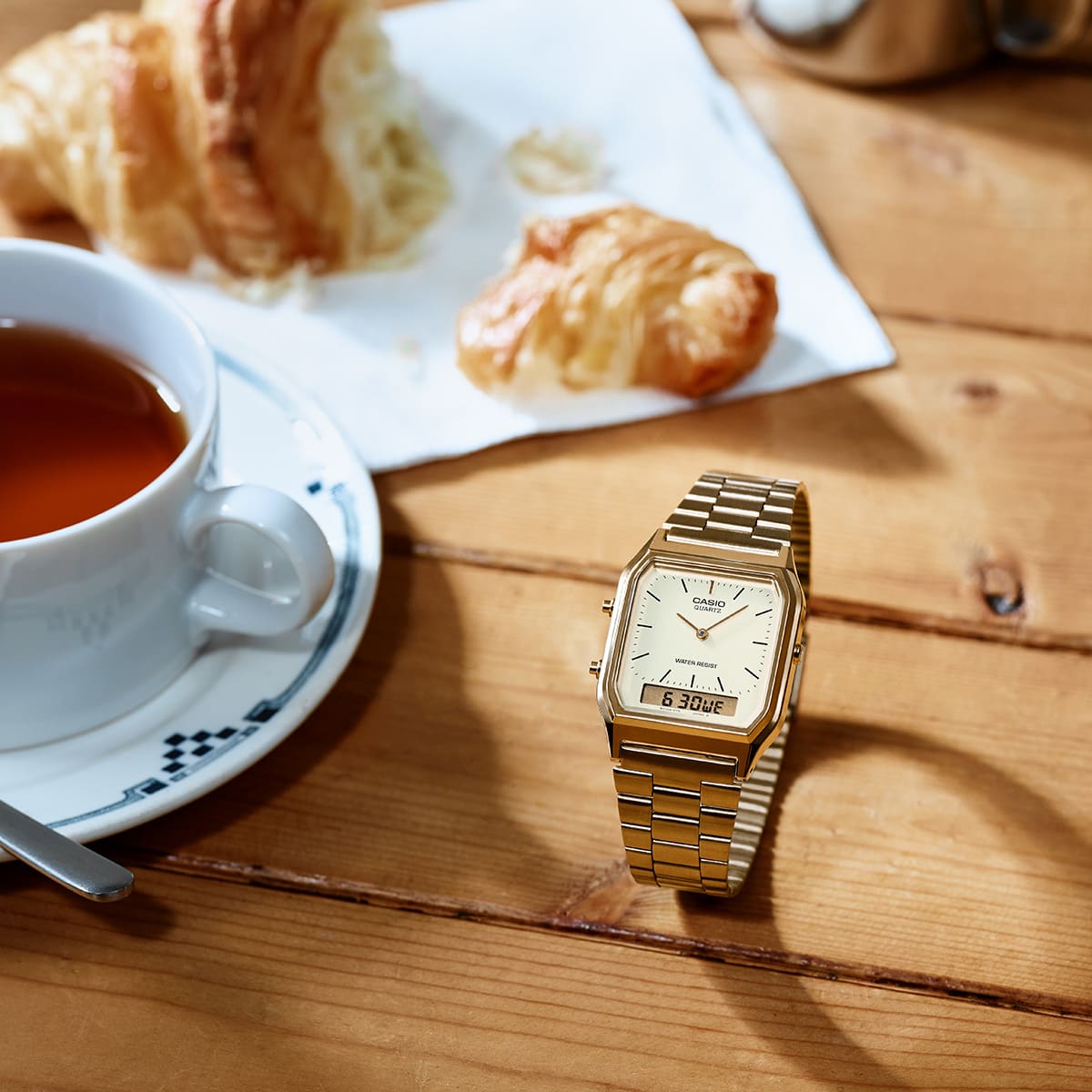 O relógio de pulso analógico dourado AQ-230 da CASIO numa mesa de madeira junto a uma chávena de chá e um croissant