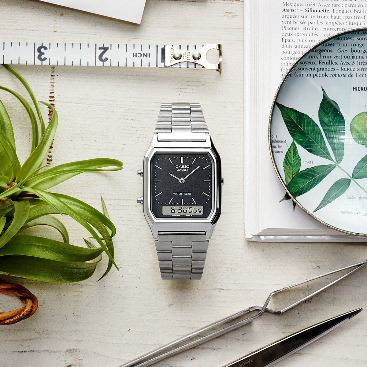 O relógio de pulso AQ-230A-1DMQ da CASIO com mostrador preto e bracelete prateada em cima de uma secretária, ao lado de uma planta e de uma lupa