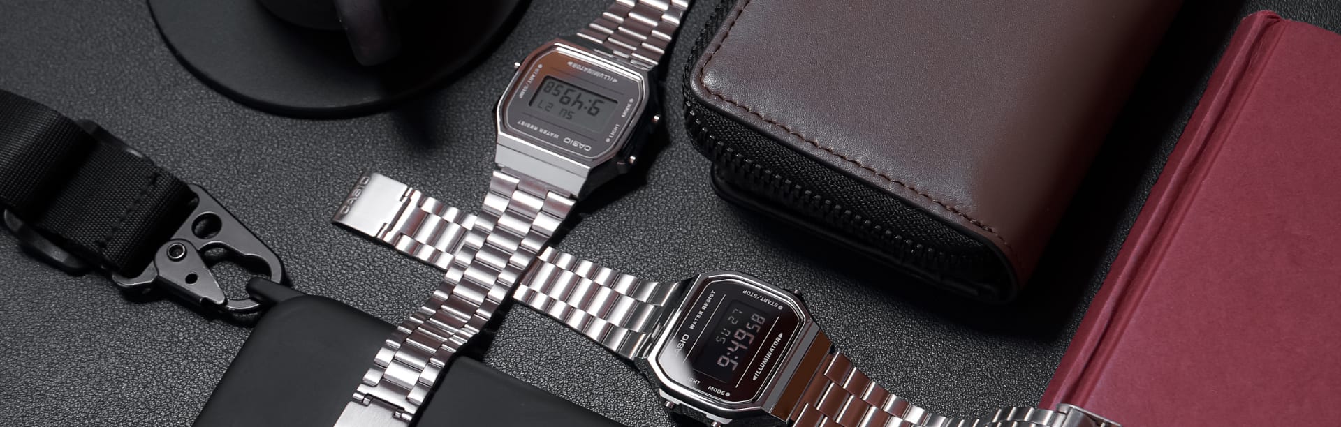 Dois relógios digitais CASIO Vintage Collection em prateado ao lado de uma bolsa, um smartphone e um notebook.