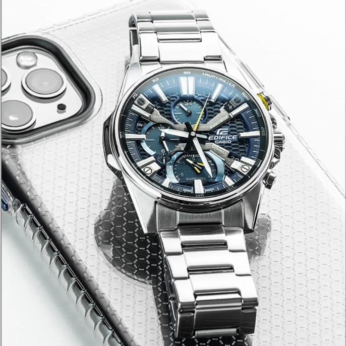 O cronógrafo EQB-1200D-2A em prateado com mostrador azul, índices brancos e detalhes amarelos sobre um iPhone com capa prateada