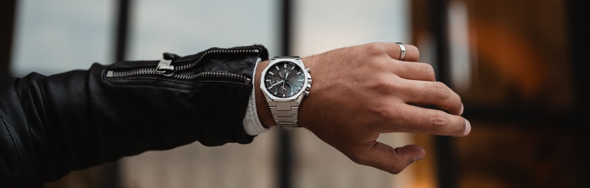 O EDIFICE Chronograph EQB-1100D-1A prateado com bracelete em aço inoxidável no pulso de um homem que veste um casaco de cabedal e estica o braço.